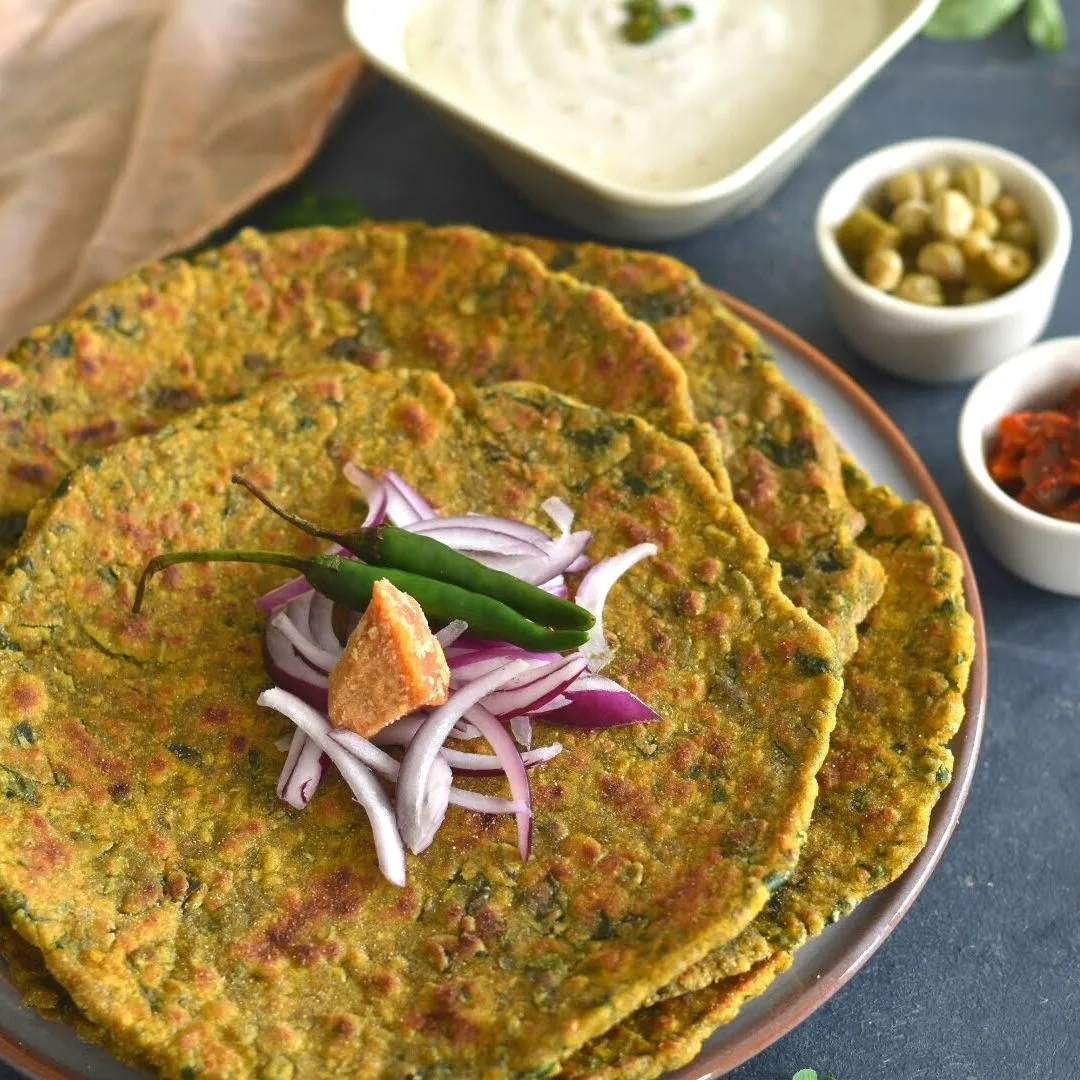 Bajra Methi Paratha served on a plate, with some cut onion, green chillies and jaggery., along with some pickles and cumin raita