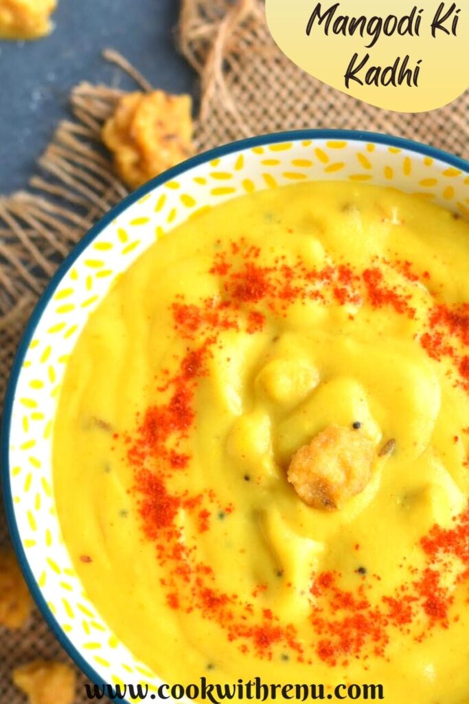 Close up look of Mangodi Ki Kadhi served in a yellow bowl. A garnish of red chilly powder and some mangodi on top. Seen in the background are some yellow mangodi
