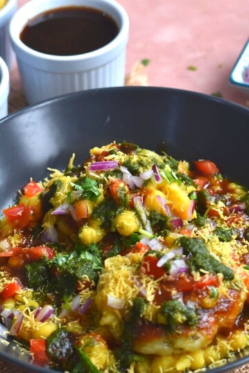Ragda Pattice served in a black plate with a generous garnish of chutney, tomato, onion and sev