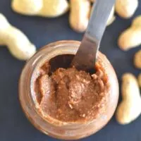 Top view of peanut butter in a glass jar with some peanuts scattered along
