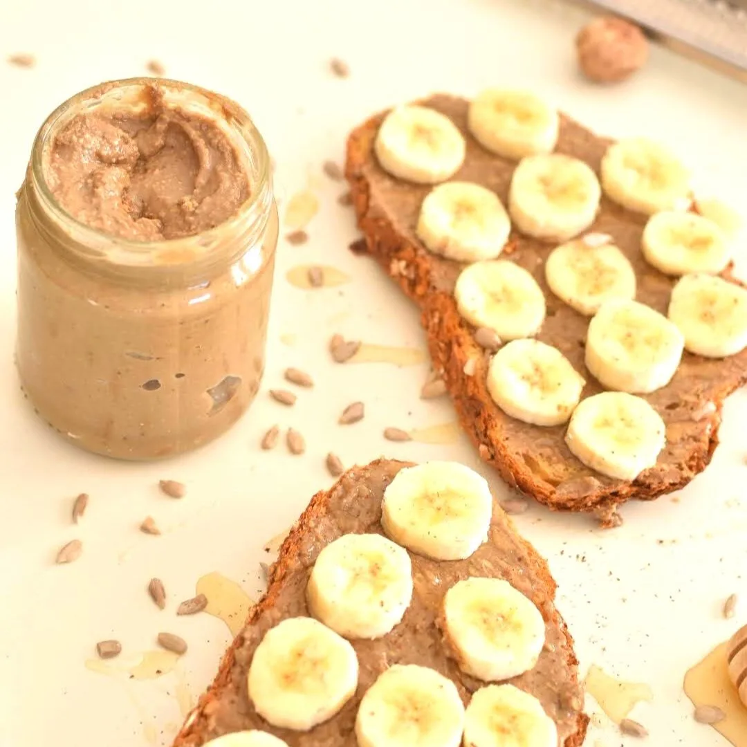 Seed Butter in glass jar along with 2 slices of toast spread with sunflower seed butter along with banana slices and a drizzle of honey and cinnamon