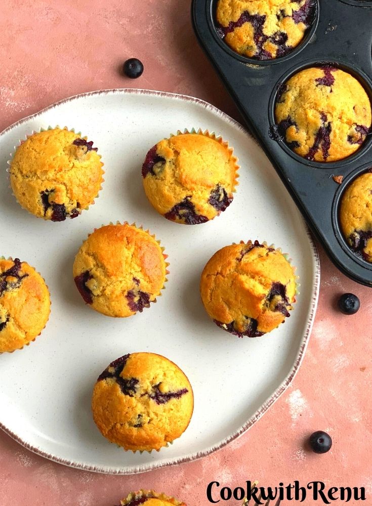 Blueberry Cornmeal Muffins arranged on a white plate with some blueberries scattered around