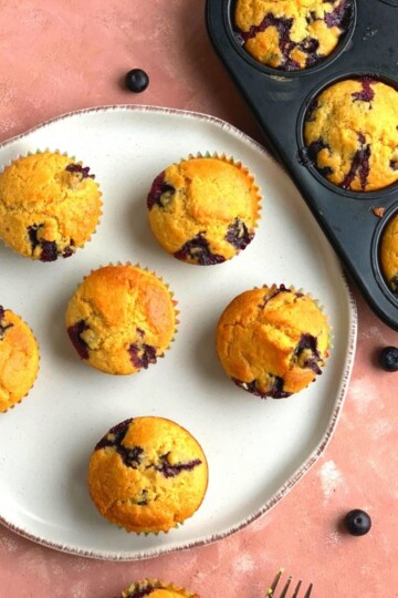 Blueberry Cornmeal Muffins arranged on a white plate with some blueberries scattered around