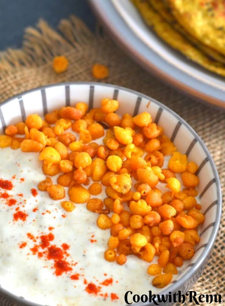 Boondi Raita presented in a bowl with a garish of boondi. Seen are some celery parathas on the side