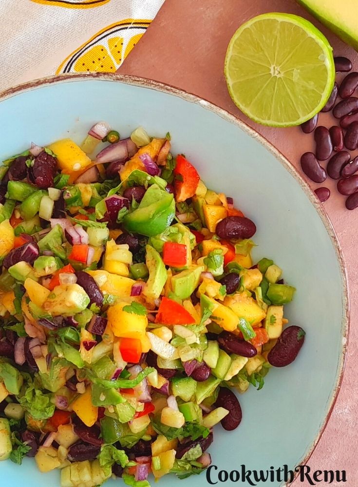 Spicy Avocado Mango Bean Salad served in a blue bowl with golden lining. A cut lime, some beans and mango is seen alongside