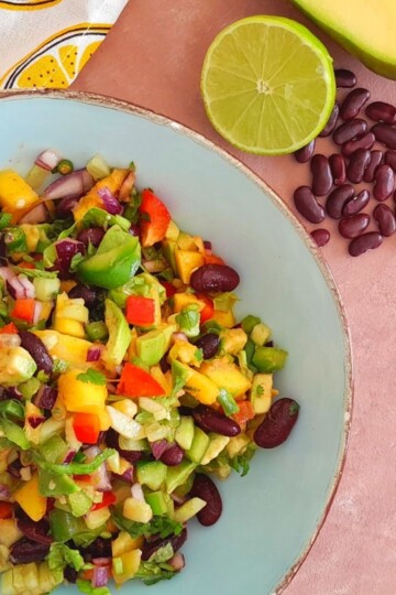 Spicy Avocado Mango Bean Salad served in a blue bowl with golden lining. A cut lime, some beans and mango is seen alongside