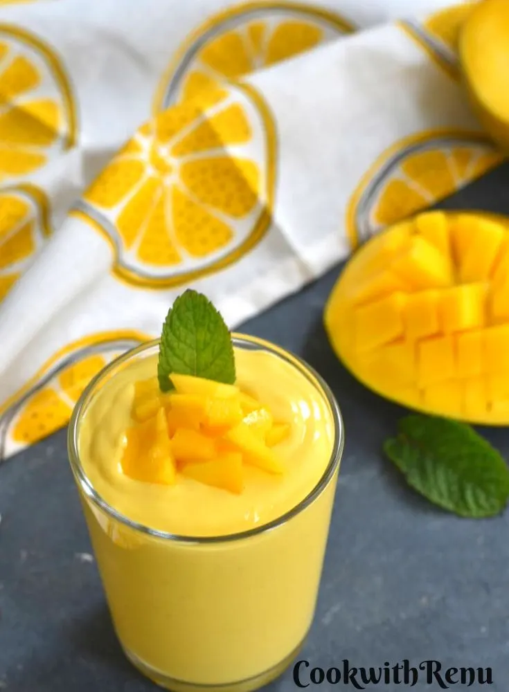 Mango Yogurt Smoothie served in a glass, with a garnish of chopped mango and mint. Seen in the background is a slice of cut mango