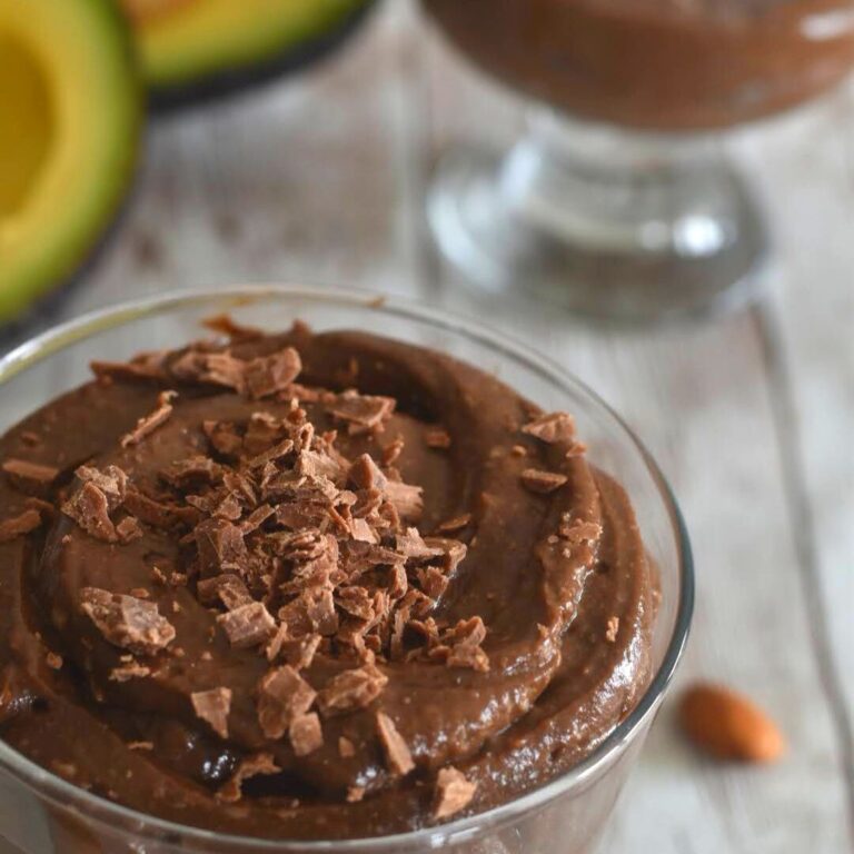 Avocado chocolate mousse served in a glass bowl with a garnish of chopped chocolate. Seen in the background are some cut avocado.