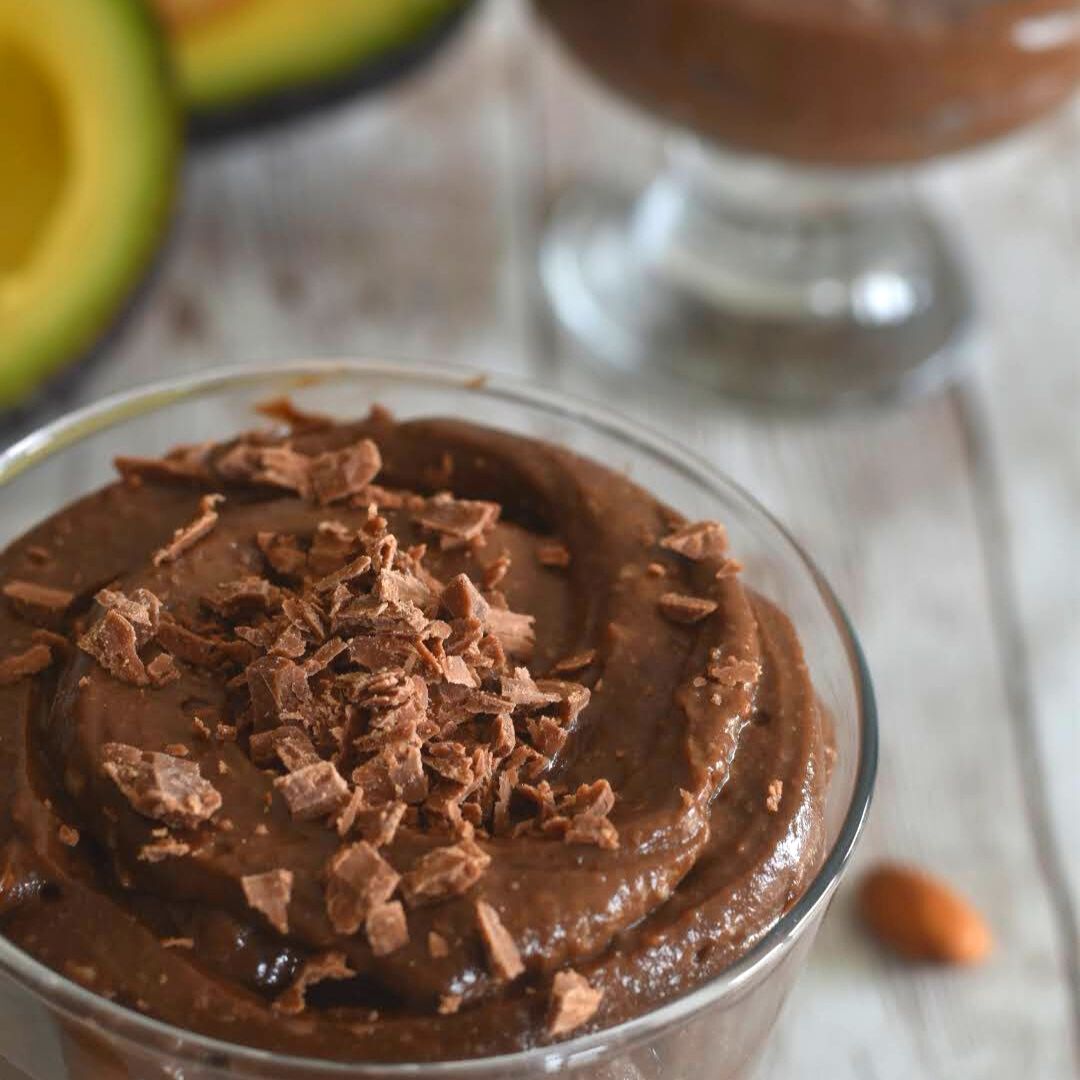 Avocado chocolate mousse served in a glass bowl with a garnish of chopped chocolate. Seen in the background are some cut avocado.