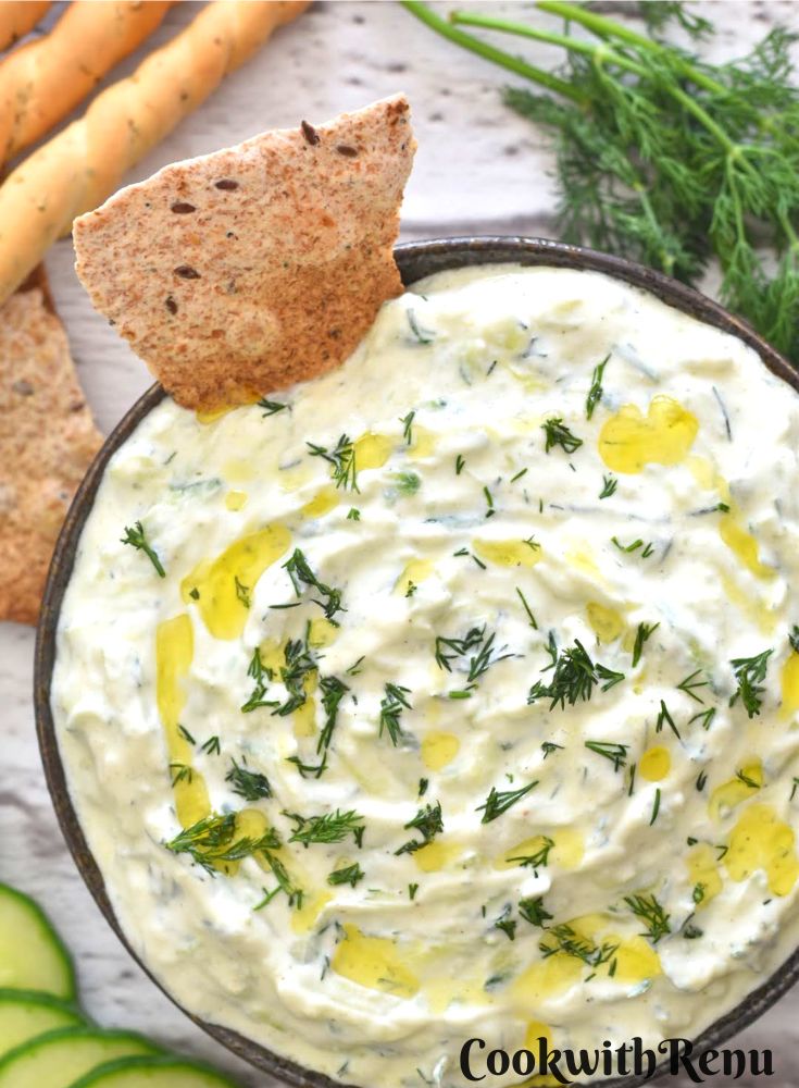 Cucumber Dill Tzatziki served in a black bowl along with some baked bread. Seen along are some fresh dill leaves and some garlic bread sticks