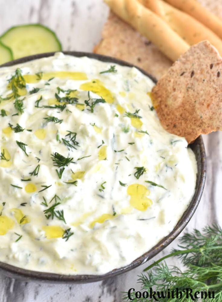 Cucumber Dill Tzatziki served in a black bowl along with some baked bread. Seen along are some fresh dill leaves, cucumber slices and some garlic bread sticks