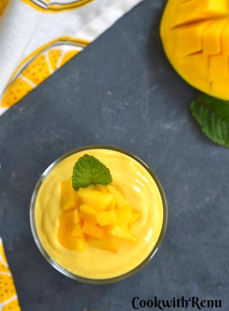Top view of Mango Yogurt Smoothie served in a glass, with a garnish of chopped mango and mint. Seen in the background is a slice of cut mango