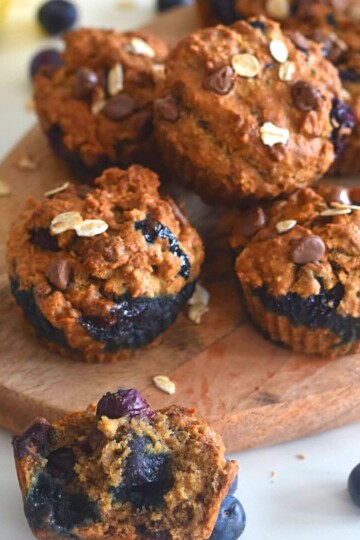 Muffins on a brown cheese board, with a close up look of half muffin in the front and few banana and blueberries in the background