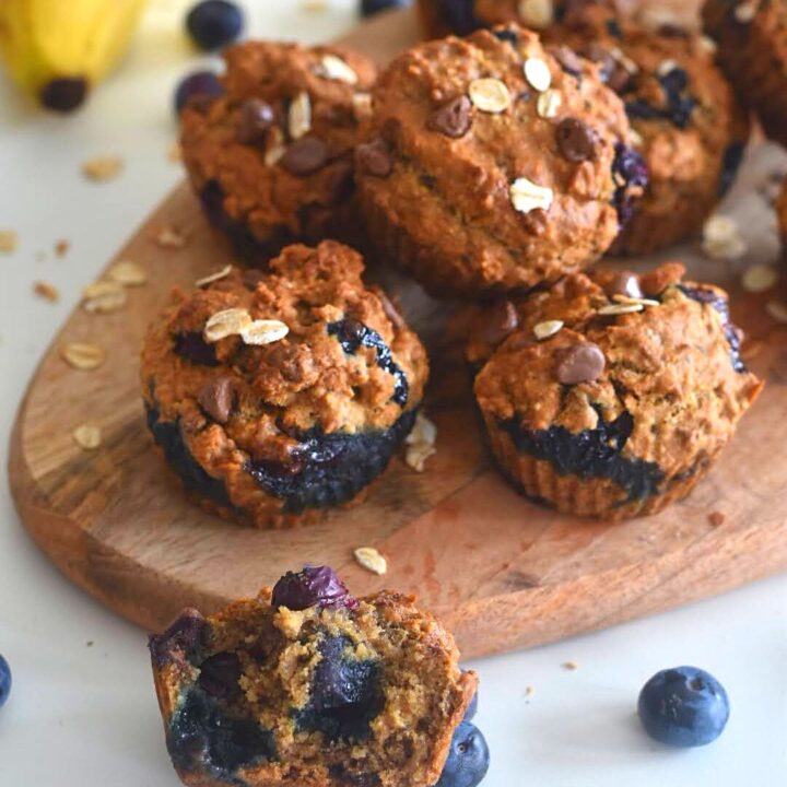 Muffins on a brown cheese board, with a close up look of half muffin in the front and few banana and blueberries in the background