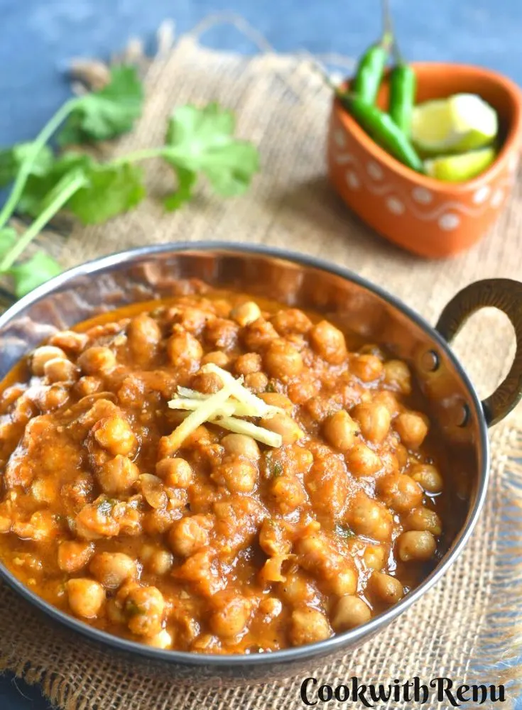 Cooked Chole in a serving kadai or wok with a garnish of ginger. Seen in the background are few coriander leaves, green chili and lemon in a brown small bowl.