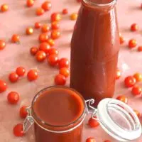 Tomato ketchup in a glass jar and a glass bottle with some cherry tomatoes lying all over.