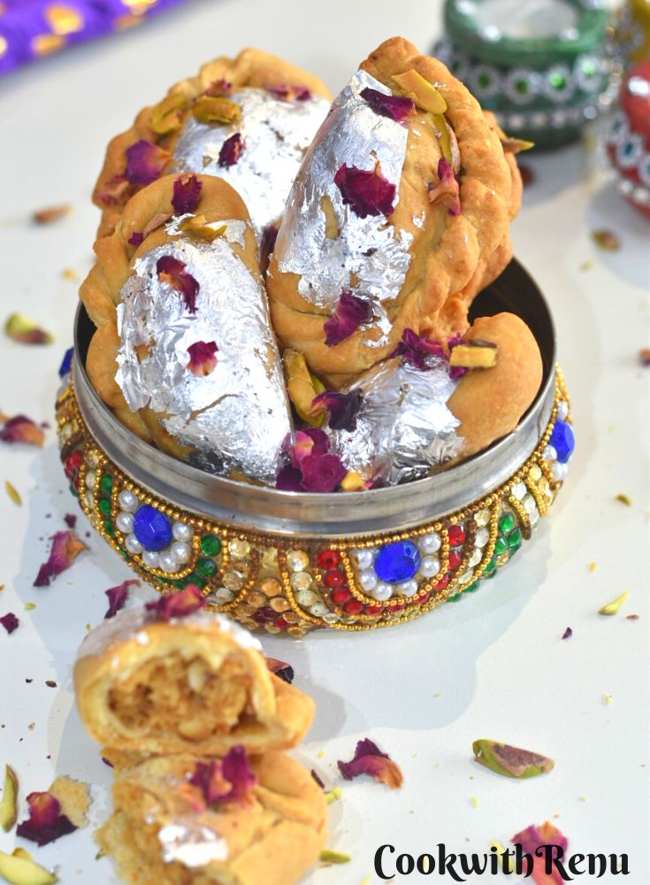 Air Fryer Mawa Gujiya presented in a designer colourful bowl. Gujiyas are garnished with edible silver leaves, rose petals and chopped pistachio. Seen in the front is a half broken gujiya and in the background some wax candle diyas.