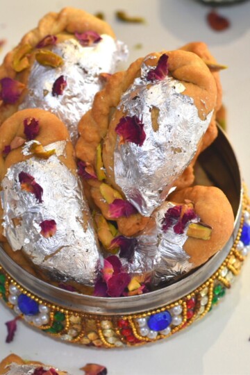 Close up look of Air Fryer Mawa Gujiya presented in a designer colourful bowl. Gujiyas are garnished with edible silver leaves, rose petals and chopped pistachio.