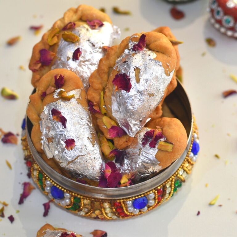 Close up look of Air Fryer Mawa Gujiya presented in a designer colourful bowl. Gujiyas are garnished with edible silver leaves, rose petals and chopped pistachio.