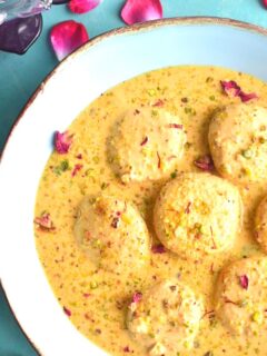 Ras malai served in a blue bowl with golden lining. Seen on the side are some rose petals.