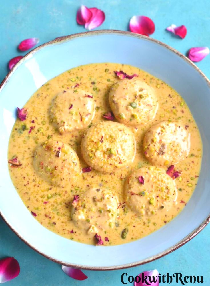 Rasmalai served in a blue bowl with golden lining. Seen on the side are some rose petals.