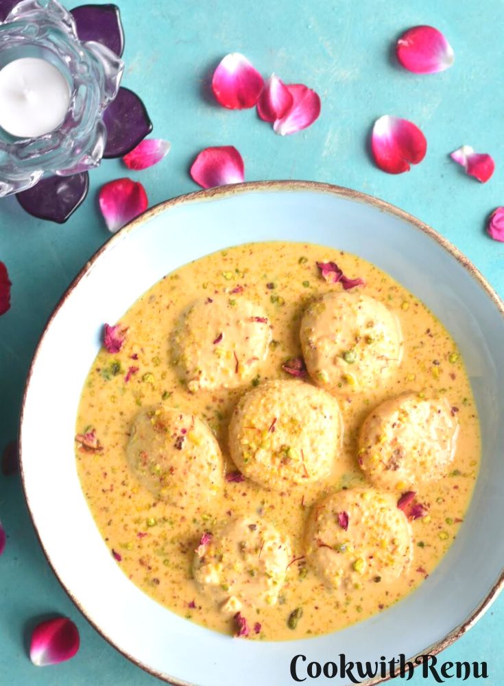 Rasmalai served in a blue bowl with golden lining. Seen on the side are some rose petals and a candle in a lotus shaped candle stand..