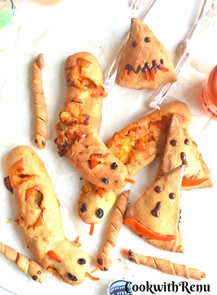 Top view of creepy Halloween Stuffed bread, witch hat and snake breadsticks arranged on a white parchment paper paper, with some festive decorations seen on the side.
