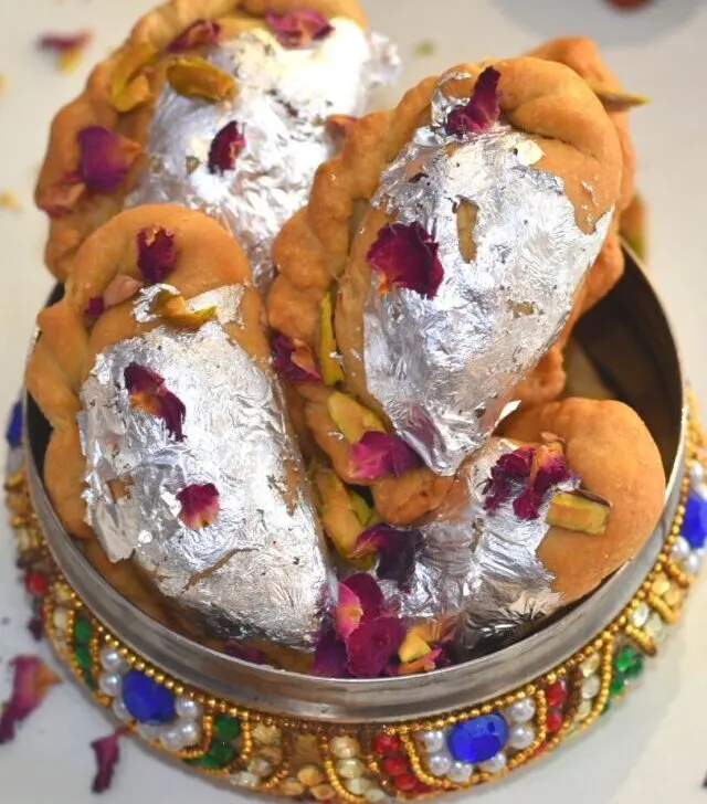 Close up look of Air Fryer Mawa Gujiya presented in a designer colourful bowl. Gujiyas are garnished with edible silver leaves, rose petals and chopped pistachio.