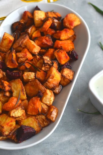 Roasted vegetables in a white rectangular tray with some yogurt chive dip. Seen in the background are some rosemary and a clean kitchen towel.