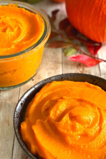Pumpkin puree in a dark brown bowl and a glass bowl, with some pumpkin seen in background and some autumn leaves.