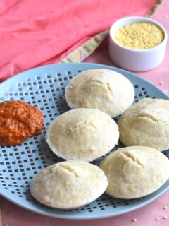 Idli served in a blue plate with Chettinad Spicy Red chili chutney. Seen in the background are some porso millet in a white bowl and a pink scarf.