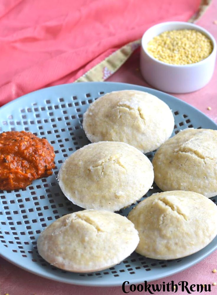 Idli served in a blue plate with Chettinad Spicy Red chili chutney. Seen in the background are some porso millet in a white bowl and a pink scarf.