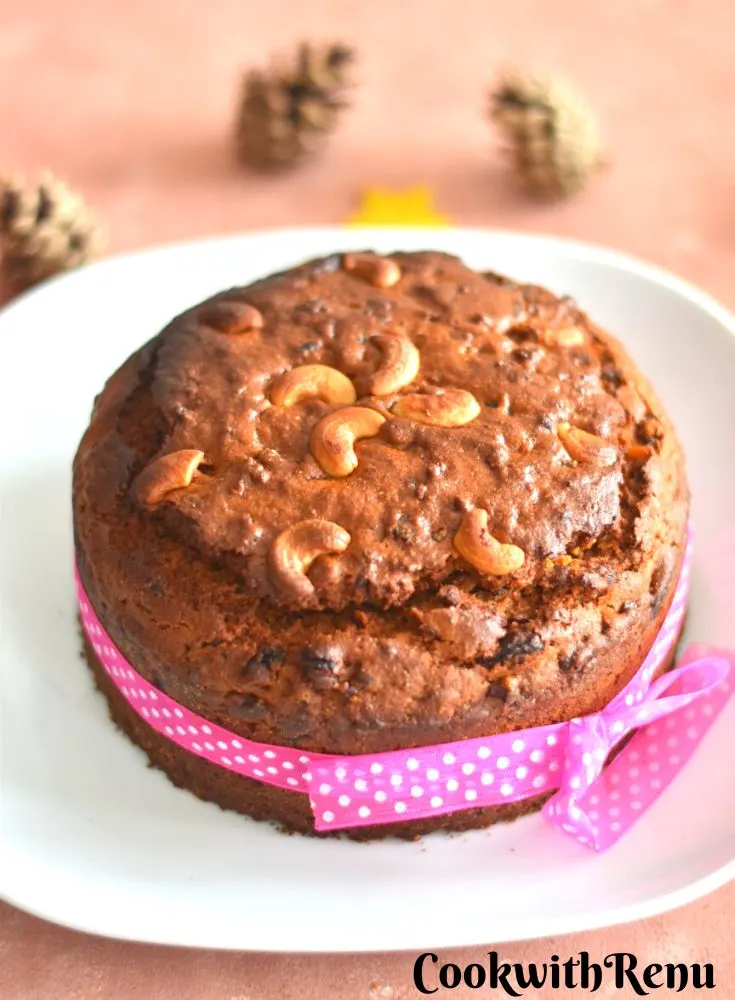 Eggless and Dairy Free Kerala Plum Christmas Cake on a white plate with some pink ribbon on the side and some Christmas decorations seen in background.