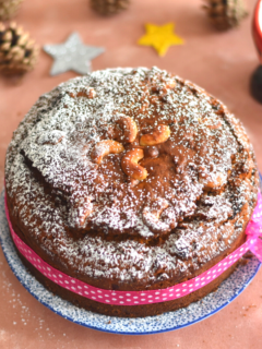 Vegan Kerala Plum Cake decorated with icing sugar and some pink ribbon. Seen in the background are some Christmas decorations.