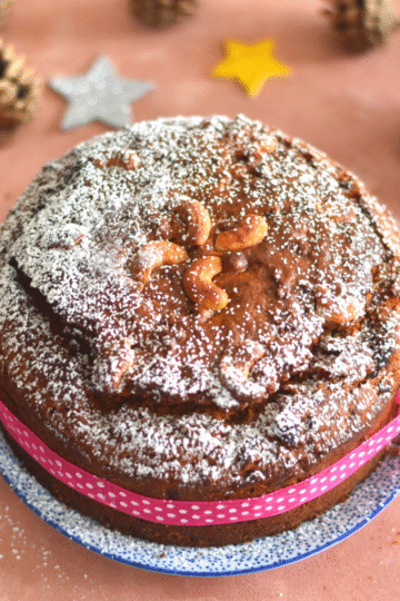 Vegan Kerala Plum Cake decorated with icing sugar and some pink ribbon. Seen in the background are some Christmas decorations.