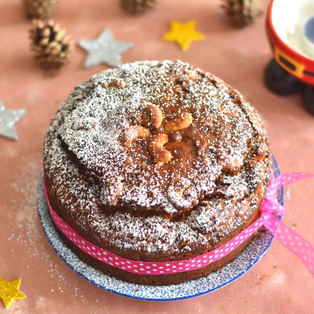 Vegan Kerala Plum Cake decorated with icing sugar and some pink ribbon. Seen in the background are some Christmas decorations.