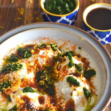 Dahi Vada or Dahi Bhalla served in a white bowl with grey lining. The vadas are topped with chutney and garnished with sev. Seen in the background is green coriander and Imli chutney.