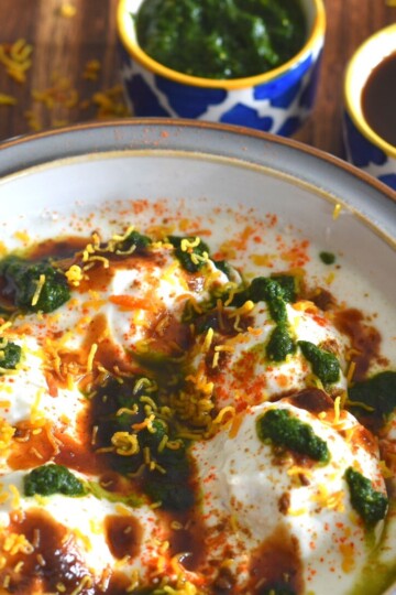 Dahi Vada or Dahi Bhalla served in a white bowl with grey lining. The vadas are topped with chutney and garnished with sev. Seen in the background is green coriander and Imli chutney.
