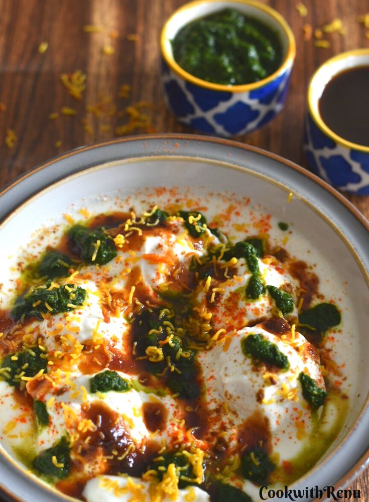 Dahi Vada or Dahi Bhalla served in a white bowl with grey lining. The vadas are topped with chutney and garnished with sev. Seen in the background is green coriander and Imli chutney.