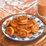 Air Fryer Whole Wheat Bhakarwadi served in a blue plate which is kept on a wooden board. Seen in the background is a glass of Indian Masala Tea.