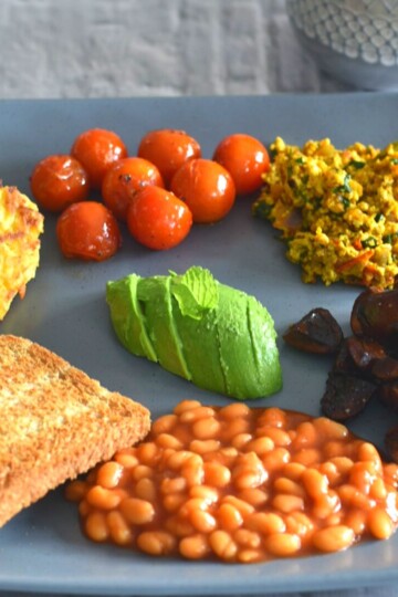 Vegan Irish Breakfast served on a grey plate along with some coffee in a grey cup. The breakfast consist of (anticlockwise from front) baked beans, sauteed mushroom, Indian style tofu bhurji, sauteed tomato, hash browns, butter and in the center avocado.