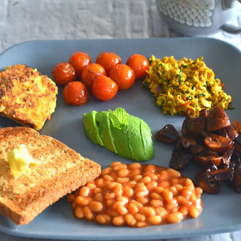 Vegan Irish Breakfast served on a grey plate along with some coffee in a grey cup. The breakfast consist of (anticlockwise from front) baked beans, sauteed mushroom, Indian style tofu bhurji, sauteed tomato, hash browns, butter and in the center avocado.