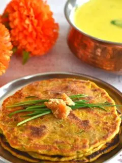 Gluten-Free Millet Chives Flatbreads served in a steel plate with some Maharashtrian Takachi kadhi or yogurt soup in a brass bowl and flowers seen in the background. A garnish of chives and jaggery is on the flatbread.