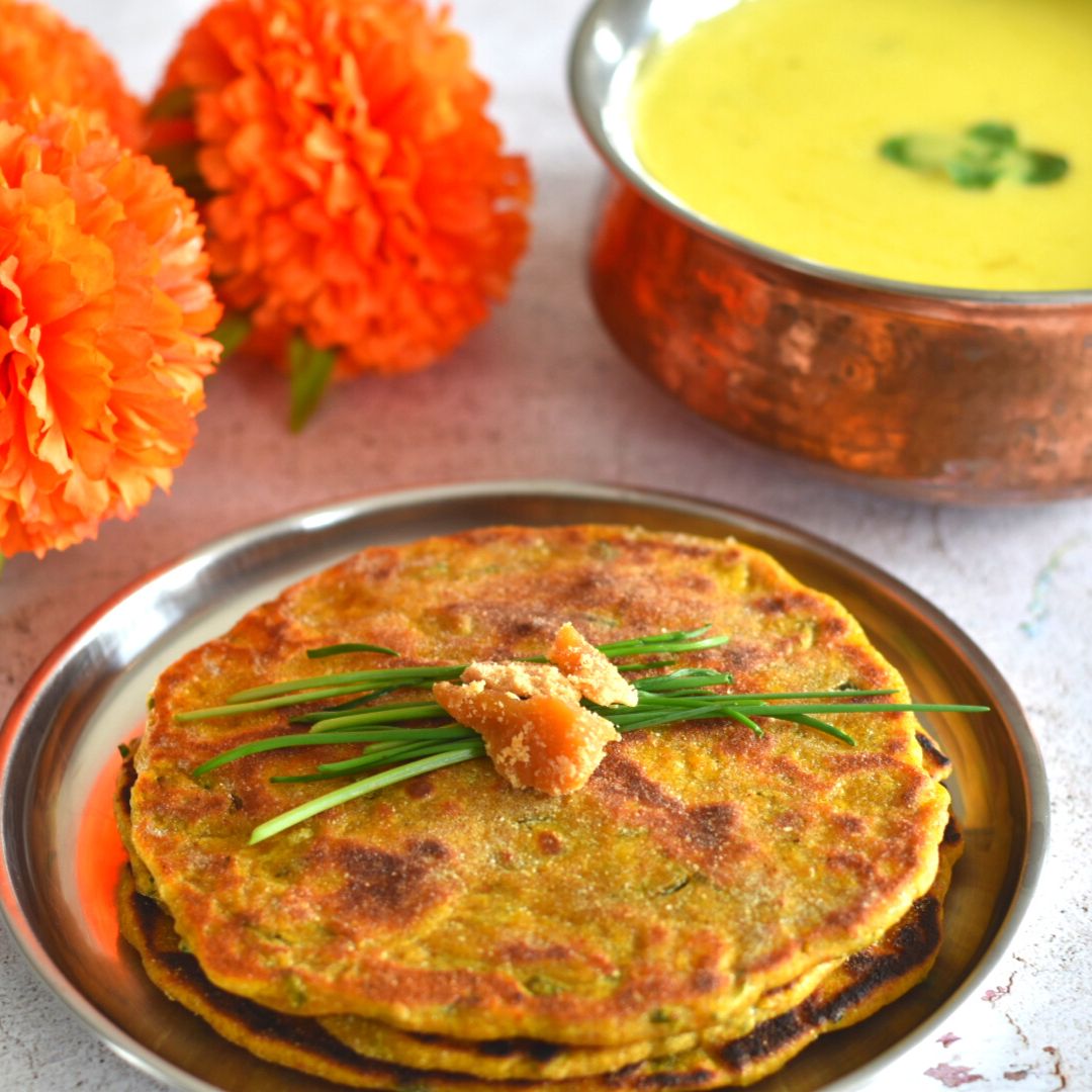 Gluten-Free Millet Chives Flatbreads served in a steel plate with some Maharashtrian Takachi kadhi or yogurt soup in a brass bowl and flowers seen in the background. A garnish of chives and jaggery is on the flatbread.