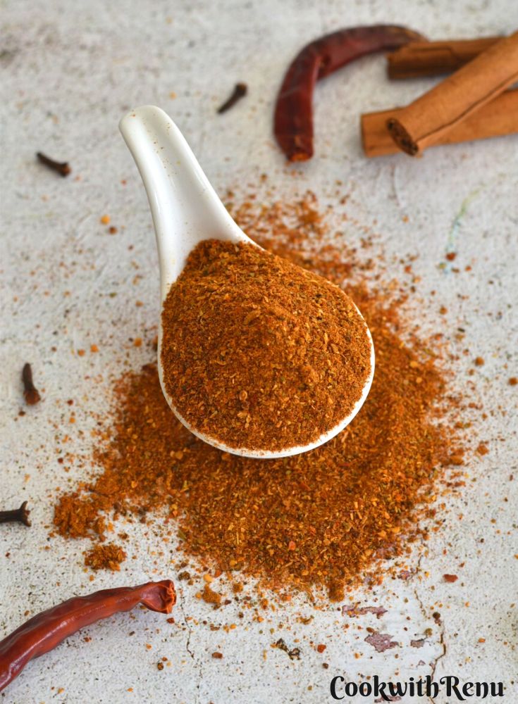 Chole masala powder in a white spoon, some scattered on the side, and a few cinnamon sticks, cloves, and red chilly on a white designer paper.