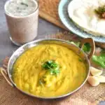 Besan Pithla served in a kadai, with garlic and coriander seen in the side along with a glass of solkadhi and rice bhakri in a plate.