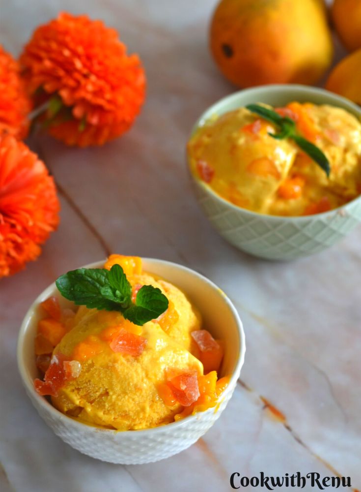 Mango Ice Cream served in a white bowls, with a garnish of tutty fruity, mango pieces and mint. Seen in the background are some fresh mangoes along with a red stole.