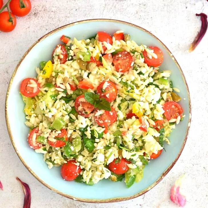 Top view of Vegetable Orzo Pasta Salad served in a blue bowl with golden lining. Seen in the background are some tomatoes and dried tulip flowers.