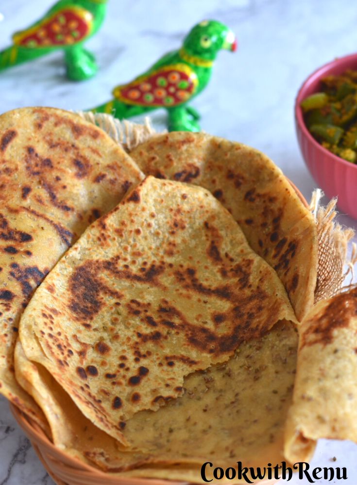 Triangular Ajwain Paratha stacked on a basket, with a green chili on it. Seen in the background is some green capsicums veg and some toys. One paratha is open and kept to show the inside stuffing.