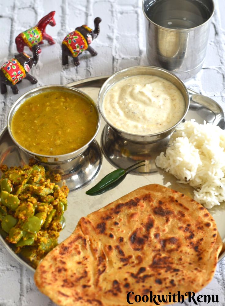 A thali with Ajwain paratha, besan simla mirchi, whole moong dal curry, dahi/yogurt, rice and a green chili. Seen besides is a glass of water and some toy elephants and horse.